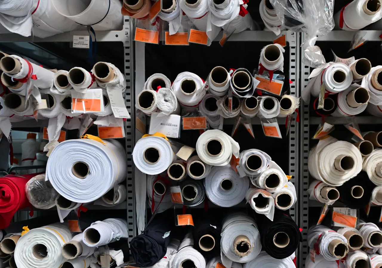 Estanterías llenas de rollos de tela de varios colores, incluyendo blanco, negro y rojo, con etiquetas que indican una fábrica de producción propia.
