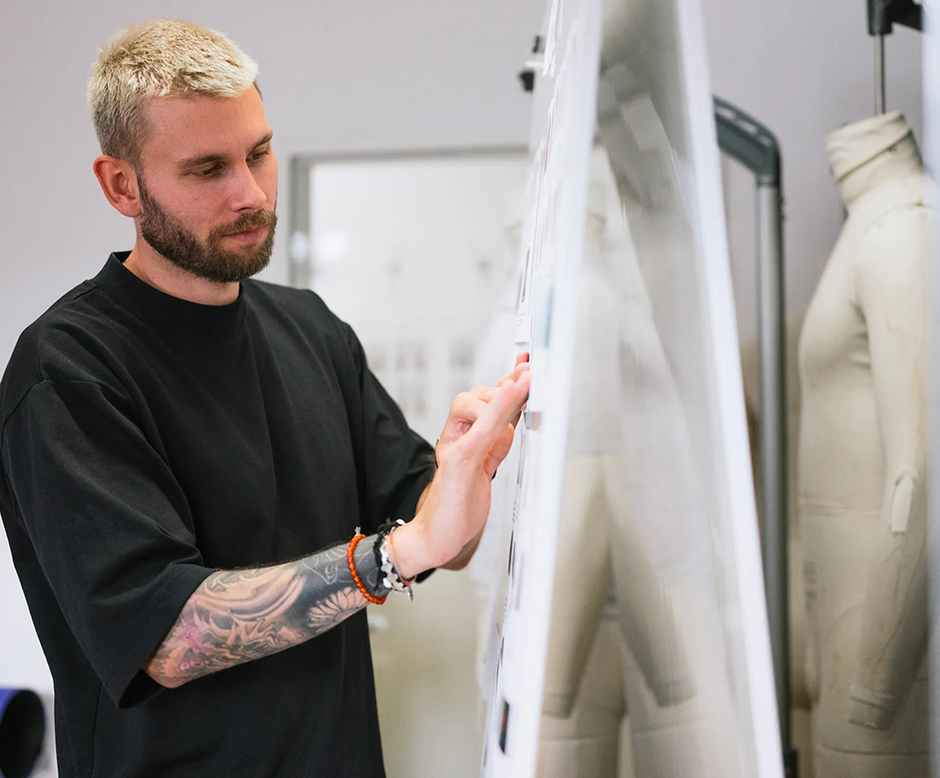 Un hombre con cabello rubio y un tatuaje en el brazo examina atentamente diseños en un tablero de anuncios en un estudio de diseño, mientras una maniquí se encuentra en el fondo.