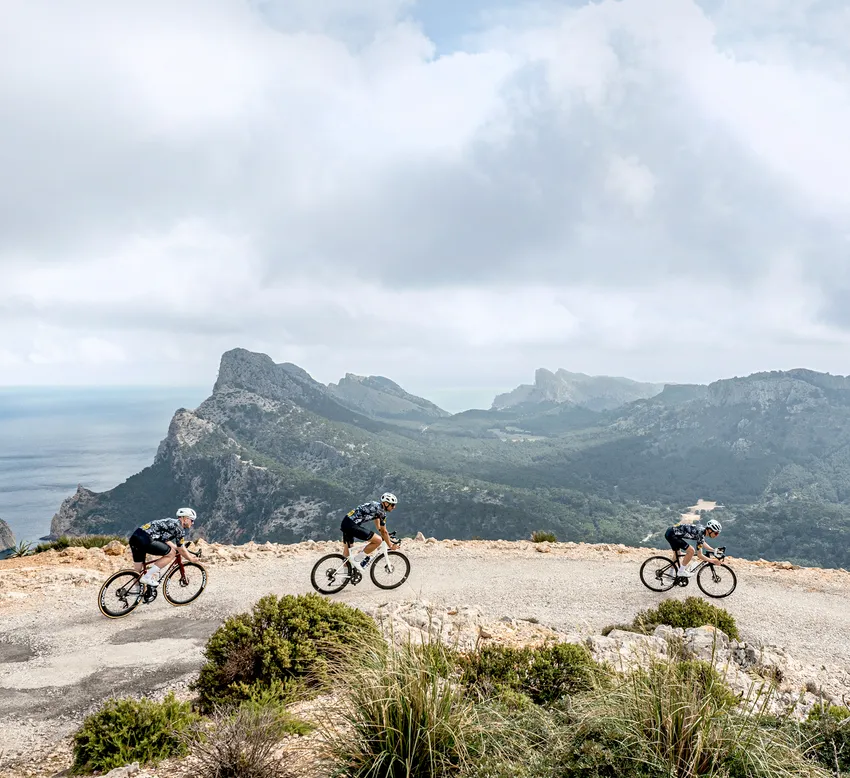 Tres ciclistas con ropa deportiva montan en una carretera a lo largo de un paisaje costero con montañas y mar al fondo.
