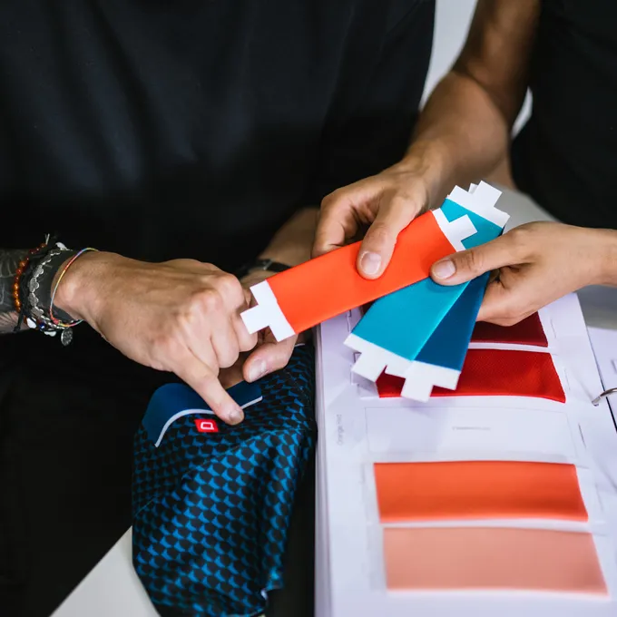 Dos personas seleccionan muestras de color para ropa deportiva personalizada, con un tejido azul con patrón y varias opciones de color visibles.